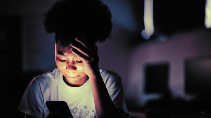 A girl sits in the dark holding her head looking at a mobile phone